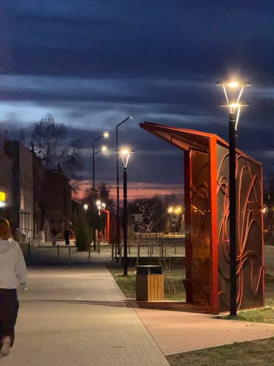 The image depicts a street scene at dusk or night in Borzya, Russia. Streetlights illuminate a pedestrian walkway, alongside which is a modern bus stop with a red frame and decorative elements on its roof. A person in light-colored clothing is visible in the lower foreground. Buildings and bare trees are seen in the background. The sky is a gradient of dark blue and hints of orange at the horizon.