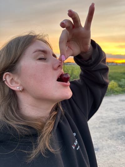 The image depicts a young person, likely female, captured from a low angle and rotated 90 degrees to the left. The subject's head and upper torso are visible, with her face turned upwards and mouth slightly open. A single red cherry, held by its stem between her thumb and forefinger, is positioned directly above her open mouth, suggesting she is about to eat it. Her fingernails are painted a light pink.
Her eyes are closed or mostly closed, conveying a sense of focus or enjoyment in the moment. Light brown, wavy hair frames her face and flows downwards. She is wearing a black hoodie, on which faint white and red text can be seen, partially spelling out "HRKA" and "RECR". Visible on her ear is a small hoop earring with a dangling silver heart charm, and another small stud earring in her earlobe.
The background, also rotated, features a vibrant, warm orange and yellow sky, indicative of either sunrise or sunset. Below the sky, some blurred green foliage and a light-colored ground or path are visible. The setting appears to be outdoors, in a natural or semi-natural environment, potentially in Горный, Russia, during twilight hours. The overall image captures a candid, serene moment of sensory pleasure.