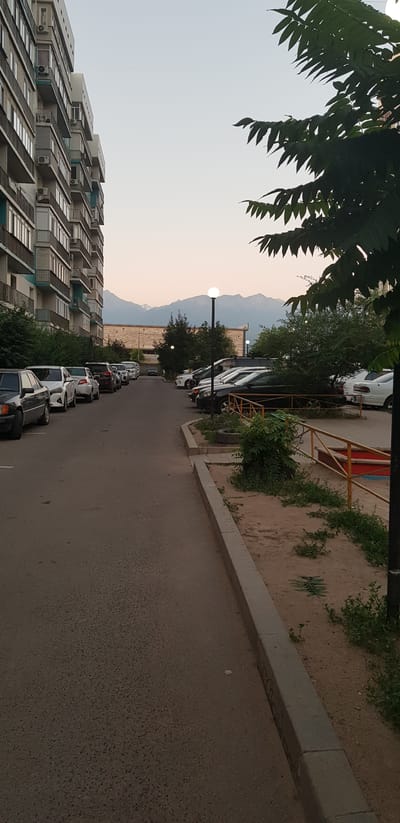 In Іргелі ауылдық округі, Kazakhstan, an outdoor scene depicts a residential area with apartment buildings and a street lined with parked cars. A multi-story apartment building with balconies and air conditioning units is visible on the left. On the right, several cars are parked along the street, which is bordered by a sandy area with a yellow metal fence and some vegetation. In the background, a larger building and trees are present, with a mountain range visible in the distance under a pale sky. The overall impression is one of a somewhat dated urban environment, possibly during dusk.