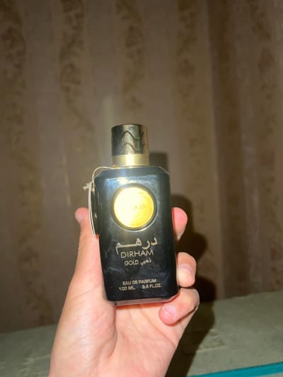 A hand holds a black perfume bottle with a gold cap. The bottle has text and a circular gold emblem on it. The background is a blurred curtain. The image does not provide any information about the city of Romitan, Uzbekistan.