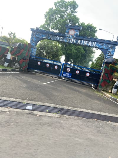 The image shows the gate to the "Pos. II  Hud Suliaman" military base, located in Kecamatan Margahayu, Indonesia (latitude: -6.978, longitude: 107.561). The gate is made of black metal bars with blue accents, topped with a blue and white archway adorned with the words "Pos. II Hud Suliaman" in white lettering. There are two palm trees on the left side of the gate and an overhead street lamp to the right. There is a "RISKALI PENGHINDUK 200M" sign on the right side of the gate, indicating a 200-meter perimeter. The gate is closed, and there is a black and white striped curb in front of the gate. There is also a white piece of trash on the ground in front of the gate, off to the left. The ground is paved in grey asphalt, and there is a small, unidentifiable object in the distance.  There are no people or vehicles in the image.