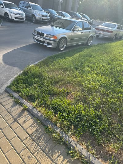 The image displays a row of parked cars along a grassy verge next to a sidewalk made of interlocking bricks. The scene appears to be a residential or commercial area in Koltsovo, Russia, given the license plates. The time of day is late afternoon, indicated by the long shadows cast by the grass and trees onto the sidewalk. The weather seems pleasant, with bright sunlight illuminating the scene.
The primary objects are several cars, including a silver BMW, a silver Toyota Land Cruiser Prado, and a white Citroën. The BMW has a license plate that reads "P498MC 154". The Toyota Land Cruiser Prado has a license plate "E921CP 154". The Citroën's license plate is partially visible as "X377OY 154". The cars are parked on an asphalt surface.
The setting is outdoors, with a neatly manicured lawn bordering a paved walkway. The perspective is from a slightly elevated angle, looking down at the cars and the surroundings. No people are visible in the frame, and there are no obvious signs of any specific event taking place; it appears to be a typical day with cars parked. The overall mood is peaceful and ordinary.