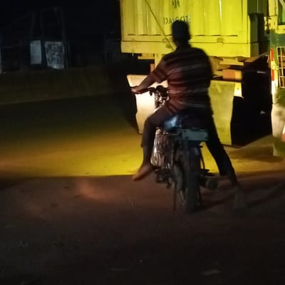 The image depicts a nighttime scene in Edopa, Nigeria, featuring an individual positioned on a motorcycle. The person, viewed from the rear, wears a horizontally striped short-sleeved shirt, dark trousers, and a dark cap. One bare foot is visible near the ground, suggesting the motorcycle is stationary or moving slowly. The motorcycle itself appears to be a standard utility model, dark-colored with visible chrome elements on its engine block and exhaust.
Behind the motorcycle, a large, bright yellow shipping container or truck body dominates the right background. The text "DAGOTE" and an emblem are partially visible on its side. A prominent yellow light source, originating from the right, illuminates the container and creates a distinct yellow patch on the dark ground directly in front of the motorcycle. The immediate foreground is dark, indicating an unlit road surface. The far background is largely indistinct, with faint, dark structures barely discernible against the night sky.