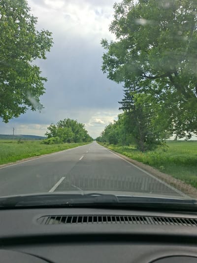 View from inside a vehicle traveling on a straight, two-lane road, lined with trees. The road surface is gray with a white line marking the edge of the right lane. Green foliage borders the road. The sky is cloudy. The vehicle's dashboard is visible on the right side of the image. The location is in Pobeda, Bulgaria.