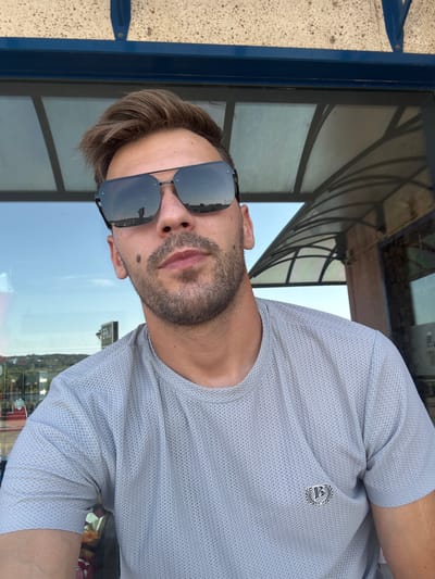 The photograph is a selfie of a light-skinned man with short, brown hair and a beard, wearing sunglasses and a light grey t-shirt with a small, circular emblem on the left chest. The man is positioned in the foreground, with a blurred background that appears to be an outdoor setting with a reflective surface, possibly a window or glass enclosure, showing a sky and some structures. A portion of a building with a textured beige facade and a dark blue trim is visible at the top of the image. The overall lighting suggests it is daytime. The man's expression is neutral, and he is looking directly at the camera.