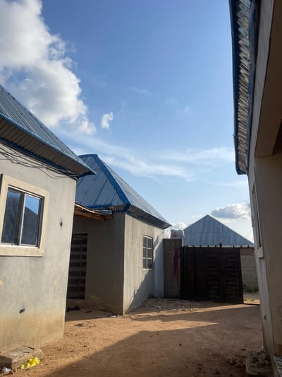 The image shows an exterior view of several residential buildings under construction in Pantinapu, Nigeria. The foreground is dominated by a dirt path leading between two buildings. To the left, a grey-walled structure with a blue corrugated metal roof features a white-framed window reflecting the sky. To the right, another grey-walled building with a similar roof is visible, partially obscured by the foreground building. Beyond these, a large, dark metal gate stands open, revealing a grey concrete wall behind it. Further in the background, another building with a peaked, blue corrugated metal roof is visible. The sky is blue with scattered white clouds. The lighting suggests it is daytime, possibly late afternoon, given the shadows. There are no people visible in the image. Scattered debris, including a yellow plastic bag, a red sandal, and black shoes, are present on the ground. The overall scene depicts a developing residential area.