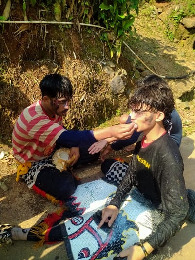 The image depicts three individuals outdoors in Gempolan, Indonesia, under bright sunlight, positioned on a dirt ground before a steep earthen bank covered in dried vegetation and some green plants.
The primary subject on the left is a male with dark hair, wearing a red and white striped t-shirt over a dark blue long-sleeved shirt, dark pants, and a black-and-white checkered fabric with red and yellow accents around his waist. His face exhibits dark, smudged markings around the eyes and cheeks. He is seated cross-legged, holding a broken coconut shell in his left hand, and extends his right hand towards the individual on the right.
The central subject, positioned to the right of the first, is a male with dark, curly hair, wearing a black long-sleeved shirt with yellow lettering (partially visible "NATOLOIN"), dark pants, and a similar black-and-white checkered fabric with dark fringes around his hips. His face also features dark, smudged markings around the eyes and cheeks. He is seated with his legs extended forward, resting upon a large, flat, woven prop adorned with red, white, and black patterns resembling a stylized animal, possibly a horse figure associated with traditional performances. His right hand rests near this prop. This individual's mouth is open, receiving an item from the extended hand of the left subject, suggesting feeding or hydration.
A third individual is partially visible behind and between the two main subjects, identifiable by hands and dark clothing, seemingly involved in the interaction or attending to the central subject. A white pipe or hose runs horizontally along the earthen bank in the background.