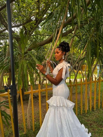 An African woman in a white gown stands in front of a yellow fence. The gown is long and flowing, with ruffled layers at the bottom and bows on the shoulders. The woman has her hair styled up in a bun and is wearing large earrings and a watch. Her hands are raised, and she appears to be holding something. Behind her, there are trees and foliage. The ground is covered with grass. A black pole, likely part of a photography setup, is visible on the left side of the image. The scene is outdoors, in a park-like setting, possibly in Barikin Gidar Kanat, Nigeria, given the woman's ethnicity and potential location.