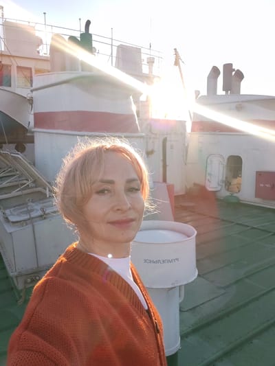 A woman with blonde hair and an orange sweater is taking a selfie on the deck of a ship. The sun is low in the sky, casting long shadows and lens flares across the image. The background shows parts of the ship's superstructure, including white and red painted sections, windows, and pipes. The deck is green and appears to be metal. The woman is smiling and looking directly at the camera. The scene suggests a maritime setting, possibly in or near Bol'shie Mkhi, Russia, given the context.