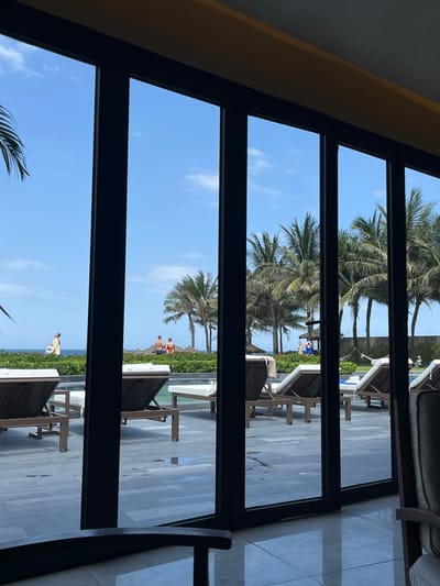 The image depicts an outdoor tropical resort scene viewed from inside a building in Cam Lâm, Vietnam, through multiple black-framed glass doors.

In the immediate foreground, a reflective light-colored tiled floor is visible, along with the dark, curved arm of an unseen chair on the left and a portion of a cushioned, upholstered chair on the right, indicating an interior space.

Beyond the glass doors, a grey-toned paved pool deck extends outwards, showing several wet spots. Numerous dark brown lounge chairs with white cushions are arranged on the deck, some facing away, some angled towards the left. A blue item, possibly a towel, rests on one chair. The edge of a swimming pool is partially visible behind the lounge chairs.

Further back, a vibrant green hedge or strip of low vegetation acts as a barrier. Behind this, a series of tall palm trees with green fronds are scattered across the midground. The clear blue sky with sparse white clouds dominates the upper background, meeting a calm, blue ocean at the horizon line.

Several individuals are present in the outdoor area. On the far left, an individual in light clothing, possibly holding a yellow towel, walks near the ocean's edge. To their right, two individuals, one male in blue shorts and one female in a red swimsuit, stand near a small thatched-roof structure, facing the ocean. Further right, behind a cluster of palm trees, two other individuals, possibly females in light tops and blue bottoms, are visible near another small structure. Another person is partially visible further right, near a white hammock strung between two posts. All individuals appear engaged in leisure activities in a bright, sunny environment.
