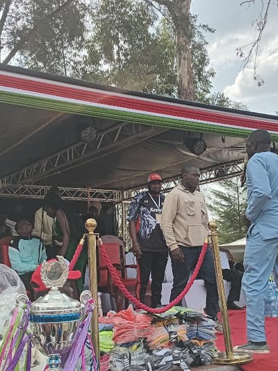 The image depicts an outdoor event taking place on a temporary stage in Manda-Shivanga ward, Kenya. The stage features a metal truss structure with two visible stage lights and a canopy. Its front is adorned with black, red, green, and white fabric, indicative of the Kenyan flag. A red carpet covers the stage floor.
In the foreground, a silver trophy with blue accents and colorful ribbons (purple, green, white) is prominently displayed, partially covered by clear plastic sheeting. Two golden stanchions linked by a red velvet rope form a barrier. Behind this barrier, a substantial pile of individually packaged clothing items, including t-shirts and socks in various colors (orange, green, blue, black, white), is arranged on the floor. Several plastic water bottles are also visible on the right foreground.
On the stage, multiple individuals are present. On the left, a man wearing a light blue and black jacket is seated on a red chair, looking upwards. Beside him, a woman with long dark hair, dressed in a dark green garment, stands. Towards the center-left, a man wearing a black t-shirt with a "Kenya" graphic and a red baseball cap stands, displaying a lanyard with a badge. Adjacent to him, another man in a beige hooded sweatshirt and dark trousers also wears a lanyard. On the far right of the stage, a man dressed in a light blue two-piece garment holds a microphone and is actively speaking. The background shows tall trees under a partly cloudy sky.
