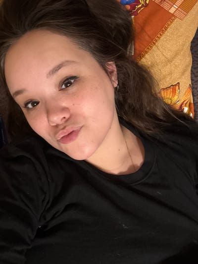 A close-up, sideways shot features a young woman lying down. She has dark brown hair that falls around her face, and her skin is lightly freckled. Her eyes are dark and looking towards the camera, with dark eyeliner accentuating them. Her lips are slightly parted, and she has a gentle, relaxed expression. She is wearing a black long-sleeved shirt, and a thin gold necklace is visible around her neck. The background shows a textured, orange and yellow patterned pillow with butterfly designs, and a dark surface. The lighting appears to be indoor, with no indication of the time of day or weather. There is no visible text in the image.