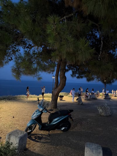The image depicts a dusk scene in Kassandra Municipal Unit, Greece, with a view of the Aegean Sea. In the foreground, a teal-colored scooter is parked on a patch of dry grass and dirt, with a large, rough-hewn stone block to its left. Beyond the scooter, a group of people are gathered on a slightly elevated area overlooking the sea. Several individuals are standing or sitting on rocks and a wooden bench, some appearing to be engaged in conversation or enjoying the view. A lamppost with a warm light is visible near a tree trunk on the right side of the frame. The sea stretches out to the horizon, with distant lights indicating a town or settlement. The sky is a deep twilight blue, with the branches of pine trees, laden with cones, forming a canopy over the scene. The overall atmosphere is one of a relaxed evening gathering in a coastal location.