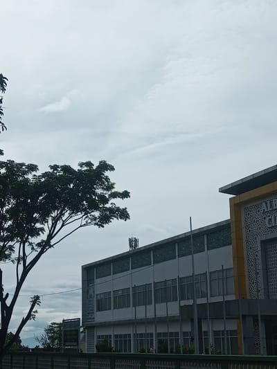 A building with multiple windows sits under a cloudy sky. Part of a tree obscures a portion of the left side of the building. On the building's roof, a small cellular tower is visible. To the right, a second building with a decorative yellow corner is present. A green fence runs across the bottom of the frame.