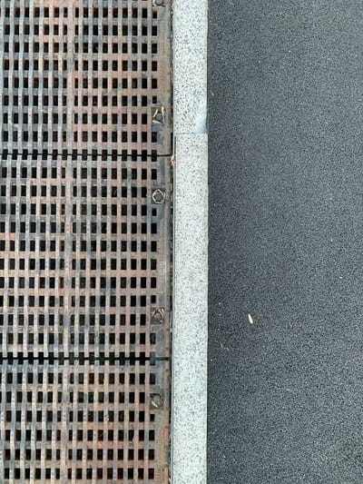 The image is a top-down view of a city sidewalk in Nizhnevartovsk, Russia. On the left is a weathered metal storm drain grate with a grid pattern of rectangular openings. To the right of the grate is a strip of granite curbing, and beyond that is a dark gray asphalt surface. There are no people or visible animals in the frame. The scene appears to be an ordinary urban environment, possibly taken on a dry day as there is no visible moisture.