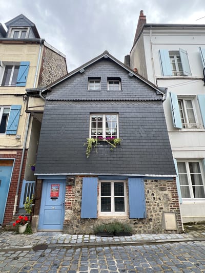 The image depicts a narrow street in the city of Honfleur, France, characterized by its cobblestone pavement and the presence of charming, historic buildings. The central focus is a tall, slender, multi-story house with a dark gray slate roof, a stone and brick foundation, and a bright blue door on the ground floor. This house features several windows, some with classic wooden shutters painted in a pale blue. Green ivy and pink flowers adorn a window box on the first floor. To the left of this house, a portion of a pale yellow building is visible, also with blue shutters. To the right, a white building rises, with more pale blue shutters and windows. The sky is overcast, suggesting a cloudy day. The street appears to be a pedestrian area, with no vehicles or people immediately visible in the frame. The overall impression is one of quaint, old-world European architecture.