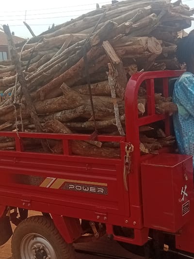 A red three-wheeled utility vehicle, likely a cargo tricycle branded "POWER," is heavily loaded with a substantial quantity of cut firewood logs in Ayangba, Nigeria. The logs, varying in diameter and length, are piled high, exceeding the height of the red cargo bed rails, and appear to be secured with dark straps. A portion of a person wearing a light blue patterned garment is visible on the far right, adjacent to the overloaded vehicle. The vehicle features a visible "POWER" decal with a '7' symbol and a hexagonal pattern on its side. A red metal toolbox, affixed to the rear right of the cargo bed, displays a white decal depicting a crossed wrench and spanner, with "A WARNING" text below. The immediate ground appears unpaved. In the blurred background, partial structures and overhead wires are discernible under a bright, possibly overcast, sky.
