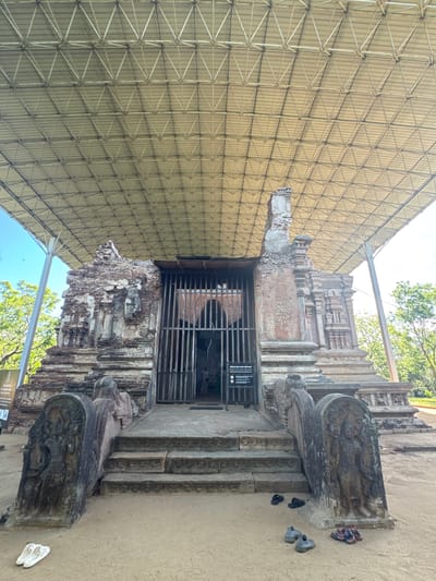 The image depicts the entrance to a historical site, likely a temple or religious building, in Polonnaruwa, Sri Lanka. The structure is made of aged brick and stone, partially covered by a large, protective metal roof structure. The entrance is gated with metal bars. Stone steps lead up to the entrance, flanked by carved stone elements. Several pairs of shoes are scattered on the ground near the steps. The surrounding environment appears to be sandy. Trees are visible in the background, suggesting an outdoor setting.