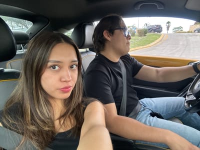 Two individuals are seated in a yellow car, positioned on a paved road with a slight incline, suggesting a street within a residential area or park. The driver, a male with dark hair and glasses, is looking towards the right side of the frame, his right hand on the steering wheel, indicating he is actively driving. He is wearing a black t-shirt and blue jeans. The passenger, a female with long, layered brown hair, is in the foreground, looking directly at the camera with a neutral expression. She is wearing a black top. The interior of the car shows black seats with yellow accents. Outside the car, a road curves to the right, bordered by a grassy area and some trees. In the background, other vehicles and a building with a stone facade are visible, suggesting an urban or suburban environment. The sky is overcast. This scene is located in Santa Clara Ocoyucan, Mexico.