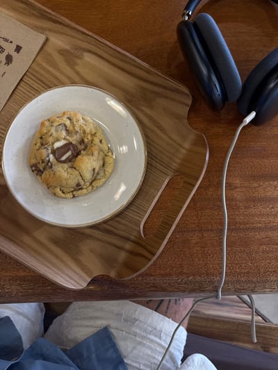 A close-up, high-angle shot shows a chocolate chip cookie on a small white plate, which rests on a wooden tray. The tray is situated on a wooden table. To the right of the tray, a pair of black headphones with a white charging cable is visible. The background is out of focus, but appears to be a blurred interior scene. No specific location in Bordeaux, France, is identifiable from the image. There are no people or animals depicted.