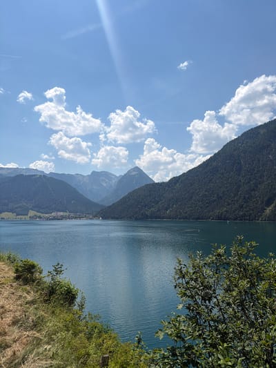 The image captures a scenic view of Lake Achensee in Pertisau, Austria. In the foreground, dry grasses and lush green bushes line a sloping embankment overlooking the vast, blue expanse of the lake. The water's surface is rippled, reflecting the bright sky.
In the mid-ground, the lake stretches out, with a few small white sailboats visible. On the far shore, a town nestled at the foot of a mountain range is visible. The mountains are covered in dense, dark green forests, with some lighter green patches indicating clearings or lighter vegetation. The peaks of the mountains are rugged and varied in shape, with some appearing sharp and others more rounded.
The sky is a clear, bright blue, dotted with fluffy white cumulus clouds. A distinct sunbeam cuts through the sky from the upper left, creating a luminous effect. The overall atmosphere is serene and picturesque, showcasing the natural beauty of the Austrian Alps.