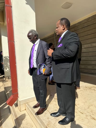 The image shows two men standing on a set of steps in front of a building. The men are both wearing suits, and the man on the left is holding a book. They are standing in front of a building in Githurai, Kenya at coordinates latitude: -1.179, longitude: 36.914. The building is white with a red-brown trim, and there is a large white rectangular ceiling fixture. The steps leading up to the building are a light grey. The man on the left is looking down, while the man on the right is looking at the man on the left. It appears to be a sunny day. There are other people in the background. The woman to the right of the man on the left is wearing a blue and white floral dress and black shoes. The man on the right is wearing a dark suit. The ground is covered in a light grey stone tile. The background is a light brown wood-look paneling. There is a red-brown rectangular pipe on the left of the image. There are shadows on the ground.