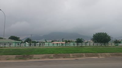 The image shows a panoramic view of a city in Nigeria, likely Dutsen Alhaji, under a cloudy sky. In the foreground, there is a road with a blurred effect, suggesting motion. Beyond the road is a strip of green grass, followed by a white picket fence. Behind the fence, several low-rise buildings with green roofs are visible, possibly part of an institution or residential area. Some of these buildings have a modern, institutional architectural style, with white walls and prominent green roofing. A dense tree stands to the right of the frame, adding to the verdant scenery. In the background, a large, hazy mountain range dominates the horizon, partially obscured by the overcast sky. The overall impression is one of a subdued, perhaps slightly damp, urban landscape.