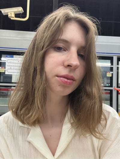 A young woman with shoulder-length light brown hair and fair skin is pictured close-up, facing forward with her head slightly tilted. She is wearing a cream-colored, textured collared shirt. Her makeup includes pink lipstick. In the background, there is a white train with blue trim. A security camera is visible in the upper left portion of the image. The scene appears to be in a subway station or a similar transit area in Uijeongbu-si, South Korea, indicated by the Korean text on a sign within the station.