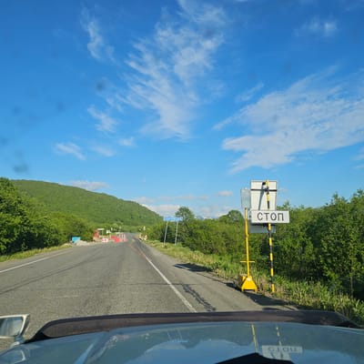 This is a media file showing a road in Dal'nii, Russia, under a bright blue sky with wispy white clouds. The camera appears to be mounted inside a vehicle, as evidenced by the reflection of the sky and trees in the car's side mirror and the car's bodywork along the right edge of the frame. The road curves to the left, bordered by lush green trees and foliage on the left and right sides.

In the middle ground, a traffic signal with a yellow and black striped barrier arm is visible, along with a sign that reads "СТОП" (STOP in Russian). Below this, a traffic light displays a green signal. Further down the road, a blue sign with white text, likely indicating a location name, is present. In the distance, a mountainous or hilly landscape covered in dense green vegetation can be seen. There are also some temporary traffic cones and barriers visible on the side of the road, suggesting possible road work or a construction zone.

The bright sky and the green landscape suggest it is daytime, likely late morning or early afternoon, with clear and pleasant weather. The scene conveys a sense of travel and journey through a scenic, natural environment. The text visible includes "СТОП" on a traffic sign and what appears to be a place name on a blue road sign, "Р. КИТАЖЕНЕЦ". There are also reflections on the car with text that is reversed and difficult to read clearly, but seems to be "ПОГРУЖЕНИЕ".