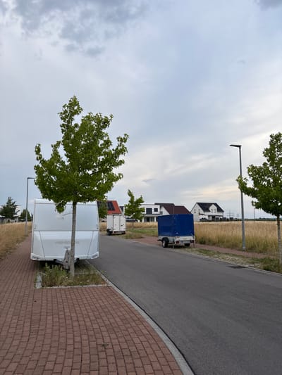 The image depicts a street scene in Bönnigheim, Germany, under an overcast sky. In the foreground, a paved sidewalk made of red bricks runs along the left side of the asphalt road. A white caravan, branded "WEINSBERG", is parked on the grass next to the sidewalk. A young tree stands beside the caravan, its branches extending upwards. Further down the road, a blue-covered trailer is parked on the grass. In the background, modern houses with dark roofs are visible, situated beyond a field of tall, dry grass. Streetlights are present along the road. The overall environment suggests a residential or semi-rural area.