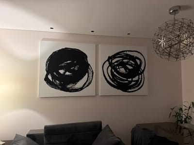 The image displays a modern interior space, likely a living room or lounge, bathed in artificial light, indicating it is evening or night in Lusail, Qatar. Two large, square canvases featuring abstract art dominate the scene, hanging side-by-side on a light beige wall. Each painting showcases bold, black, brushstroke-style circular or swirling patterns against a crisp white background.
The ceiling, painted white, is fitted with several recessed downlights that illuminate the room. A contemporary spherical chandelier with a complex metallic, geometric design hangs from the ceiling on the right side of the frame, serving as a prominent decorative element. In the lower left foreground, a portion of a dark-colored sofa or sectional is visible, with a grey pillow resting on it, suggesting a comfortable seating area. In the background on the far right, a small green plant in a pot sits on a dark table or cabinet. No people are present, and no specific activity is taking place. There is no visible text in the image. The overall aesthetic is clean, minimalist, and modern.