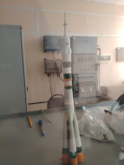A model rocket is displayed in a room with various electronic equipment mounted on the wall. The rocket is white with green and orange bands and appears to be a detailed replica. On the table in front of the rocket are a yellow pencil, a blue pen, and a paintbrush. Behind the rocket, stacked electronic units with numerous buttons, dials, and indicators are visible on the wall, suggesting a laboratory or control room setting. A large gray cabinet is to the left of the equipment. The lighting suggests it is daytime, and the overall atmosphere is one of study or experimentation. The room appears to be indoors, with plain walls and ceiling. There are no people visible in the image. Some of the electronic equipment has visible text, but it is too blurry to read. A small Russian flag is visible on the rocket.