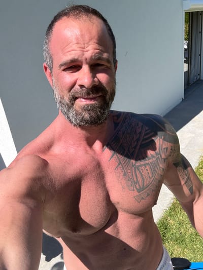 The image captures a shirtless man outdoors under bright sunlight in Marche-en-Famenne, Belgium. The man, who appears to be in his 40s or 50s, has a salt-and-pepper beard and dark hair. He is extensively tattooed with tribal-style designs covering his left shoulder and a portion of his chest. His skin shows a reddish tone, indicative of recent sun exposure or a tan. He is looking directly at the camera with a slight smile and a tilted head, suggesting a casual selfie.
The scene is brightly lit, with strong, defined shadows falling across his body, characteristic of a clear, sunny day, likely late morning or early afternoon. The background on his left consists of a plain white wall or building surface, while to his right, there's a patch of green grass adjacent to light-colored paving or concrete. Metallic structures are partially visible at the top edge of the frame, possibly part of a railing or overhead covering. A small detail of blue fabric, possibly from clothing or an accessory, is visible near his right armpit, and a white waistband is visible near his hip. No visible text is present in the image.
