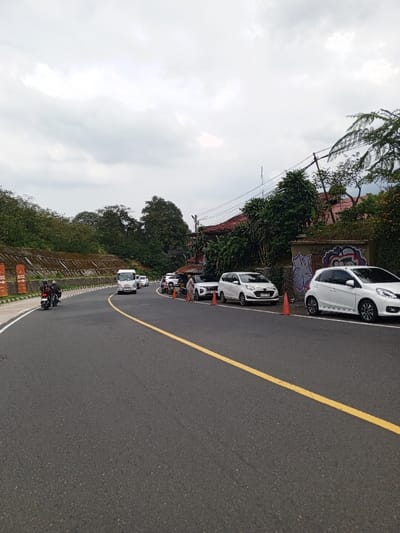 The image displays a curved asphalt road scene in Ciloto, Indonesia, under an overcast sky. The road features a solid yellow center line and a white shoulder line on its left. Traffic, including a white box truck and at least two motorcycles, is observed moving away from the viewer on the road.
On the right side of the road, several vehicles are parked parallel to the curb, including multiple white sedans/hatchbacks and a white SUV. Two orange traffic cones are positioned near these parked vehicles. Multiple individuals are standing near the parked cars; one person in a light-colored, long-sleeved garment is prominently visible near a white SUV. Behind the parked vehicles, a concrete wall exhibits graffiti in purple, black, and white. Dense green foliage, including trees and bushes, is present behind this wall, partially obscuring a building with a reddish-brown tiled roof. Overhead utility lines are visible crossing the scene on the right.
On the left side of the road, a tiered concrete retaining wall runs parallel to the road, adorned with several orange banners or flags. This wall supports a dense embankment of green trees and vegetation. The sky is uniformly light grey, indicating complete cloud cover.