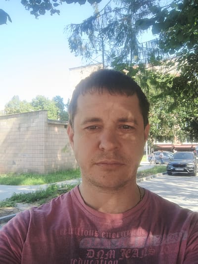 A medium shot selfie captures a Caucasian male with short, dark hair and a light stubble, looking directly at the camera. He is wearing a faded pink t-shirt with a dark graphic design on the front. The man is positioned in the foreground, with his face and upper torso filling most of the frame.
The background indicates an outdoor urban setting. To the left, a pale brick building with a tiled facade is visible, surrounded by overgrown grass and a sidewalk. Further back, trees with green foliage are present, and a section of a building with multiple windows can be seen behind them. To the right, a paved road runs horizontally. On the road, a dark-colored SUV is parked, and a silver sedan is driving away. Two individuals, a female and a male, are walking on the sidewalk in the distance, near the silver car. The sky above is a clear, bright blue. The overall lighting suggests a sunny day. The location is identified as Rivne, Ukraine.