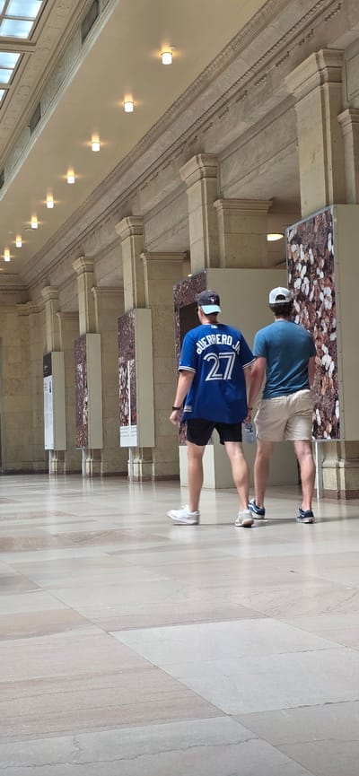 Two individuals, identified as males by their attire, walk away from the camera in an interior hallway of a large building. The person on the left wears a blue baseball jersey with "GUERRERO JR" and the number "27" in white lettering, black shorts, a dark baseball cap, and white sneakers. The person on the right wears a teal t-shirt, light-colored shorts, and dark sneakers.
The hallway features a high, arched ceiling with multiple recessed lights. The walls are constructed of light-colored stone, with ornate decorative molding running along the top. Several large display panels, featuring a close-up, abstract pattern of natural elements like leaves and nuts, are positioned along the right wall. The floor is a polished stone tile in a light, neutral color with subtle veining. The overall impression is that of a museum or a grand public building.
The image is likely taken within the Royal Ontario Museum (ROM) in Toronto, Canada, based on the distinctive architecture and the presence of large display panels.