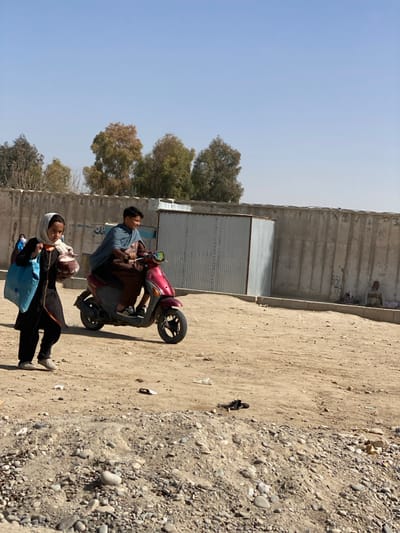 A man rides a red scooter on a dirt road with a woman walking alongside him. The woman carries a blue bag and a brown purse. In the background is a tall concrete wall, a corrugated metal structure, and several trees. The ground is rocky and dusty. The sky is clear and blue.