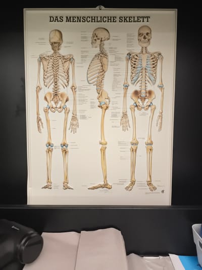 A poster depicting the human skeleton hangs on a dark wall. The poster shows three views of the skeleton: anterior, lateral, and posterior. Text labels various bones in German. Below the poster, a black surface is visible, on which lie several white paper towels. To the left, a black device with a cylindrical end is partially visible. The scene is indoors, likely in a professional or educational setting. The poster indicates it is from Troisdorf, Germany.