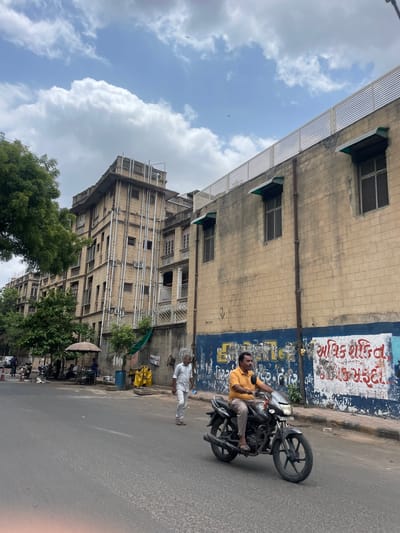 The image captures a street scene in Ahmedabad, India, with a focus on a man riding a motorcycle on a paved road. In the background, a multi-story building with a beige brick facade stands prominently. The building features several windows, some with green awnings, and external pipes running vertically along its side. A white slatted fence is visible on the rooftop. To the left of this building, another older, taller building with a similar brick construction and numerous windows is partially visible, with a large tree in the foreground. The road has some subtle textural variations. On the wall of the beige building, there is graffiti in a blue and white color scheme. Another man in a light-colored shirt and pants is walking on the sidewalk to the right of the motorcycle. Further down the street, a small roadside stall with an umbrella can be seen, with several motorcycles parked nearby. The sky above is partly cloudy with a blue hue.