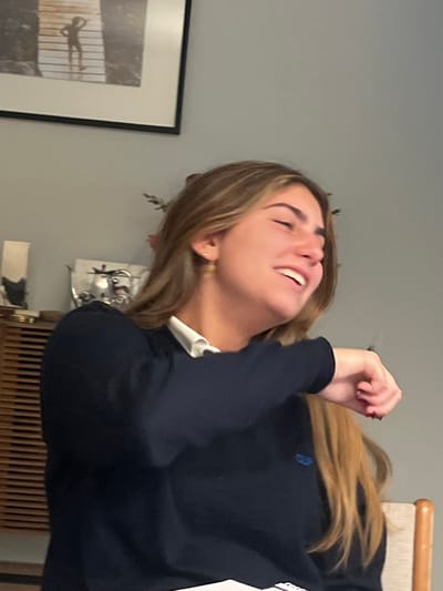 An indoor, close-up photograph features a young adult female within a domestic setting in Lordelo do Ouro e Massarelos, Portugal. The subject has light skin and long, light brown hair, captured mid-laugh or strong smile with her eyes nearly closed and mouth open. She is wearing a dark blue long-sleeved sweater over a white collared shirt; a small blue embroidered logo is visible on the sweater's left chest. Small gold-colored hoop earrings are present, and her fingernails display dark red polish. Her right arm is bent and raised, with the hand partially clenched. The background reveals a plain light gray wall. In the upper left, a black-framed picture depicts a silhouetted figure on a pier. Below it, a wooden cabinet with horizontal slat detailing holds decorative objects, including a silver-toned vessel. A portion of a light-colored wooden chair back is visible in the lower right.