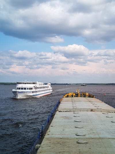 In a wide-angle, outdoor shot taken from a vessel on the Volga River near Krokhino, Russia, two boats navigate the dark, choppy water under a dynamic sky filled with cumulus clouds. In the foreground, the deck of a flat-topped barge, painted a weathered pale yellow with various circular hatches, stretches into the distance. A blue railing runs along the left edge of the barge. Further ahead, a white passenger ship, identified by the name "СЕРГЕЙ ЕСЕНИН" (Sergei Yesenin) in blue lettering on its side, cruises from left to right across the frame. It has multiple decks with windows and a prominent bridge structure. The riverbanks, lined with green trees, are visible in the background, with a few distant structures and buoys. The water is a deep blue-gray, reflecting the overcast sky. A bright orange buoy is positioned in the middle ground on the right. The overall impression is one of active riverine transport under a partly cloudy sky.