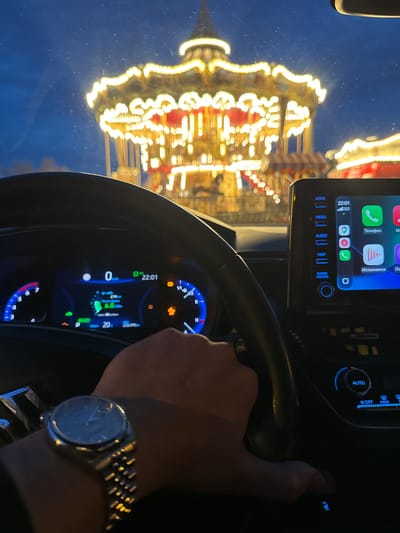 The image shows a person's hands on the steering wheel of a car at night. The dashboard is illuminated with a digital display showing the time as 22:01, a speedometer reading 0 km/h, and other vehicle information. The car's infotainment screen is visible, displaying icons for phone, messages, and navigation. Outside the car, a brightly lit carousel with yellow and red lights is visible, suggesting a festive or amusement park setting. The reflections on the windshield indicate that the car is likely parked or moving slowly past the carousel. The overall scene conveys a sense of evening leisure or travel. The visible text includes "HOME", "MENU", "AUDIO", "MAP", "POWER VOLUME", "AUTO", "OFF FRONT", "READY", and phone app names in Russian like "Телефон" (Telephone) and "Исполняется" (Playing). The time stamp "22:01" and "6.0" (possibly fuel consumption) are also displayed.