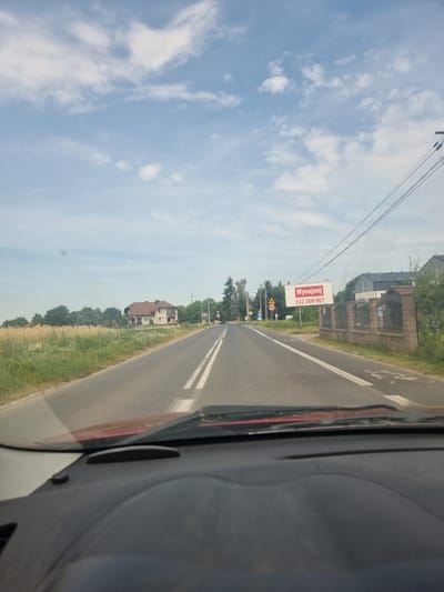 The image is taken from the perspective of someone driving a vehicle on a paved road. The road has a double white line down the center and appears to be in a rural or suburban area. To the left of the road, there is a field of tall grass and wildflowers, and further in the distance, a large, light-colored house with a brown roof. To the right of the road, there is a low stone fence with decorative metalwork, and behind it, a modern building with dark siding. A billboard is visible above the fence, featuring red text that reads "Wynajmij" and a phone number "532 300 067". Traffic signs, including a speed limit sign for 40, are also present along the roadside. The sky is blue with scattered white clouds. The image captures a daytime scene. No people are visible in the image.