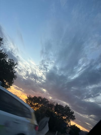 The image depicts a sunset or sunrise scene over Blizikuće, Montenegro. The sky, angled upwards from left to right, transitions from a clear, light blue at the upper left to vibrant yellows and oranges at the horizon, indicating a low sun position. A significant portion of the sky is covered by clouds: high, wispy cirrus clouds are visible against the bluer sections, while denser, darker cumulus or stratocumulus clouds dominate the right side of the frame and form a band above the horizon. These darker clouds exhibit tones of grey, purple, and blue, with patches illuminated by the low sun, creating lighter grey and white highlights.
Below the horizon, a dense line of trees, appearing as dark silhouettes, spans the lower mid-ground. Their dark foliage suggests deciduous trees. On the far right, a dark, indistinct landmass or distant hills are visible at the very bottom of the frame. In the lower left quadrant, a portion of a white or light-colored vehicle is visible, exhibiting motion blur consistent with being photographed from a moving perspective. A yellow or tan stripe runs horizontally along its side. The dark, curved shape on the extreme right edge of the image suggests the interior frame or window of the vehicle from which the photograph was captured. No individuals are present in the scene. The overall environment is an outdoor setting, likely rural or semi-urban, with natural foliage prominent against the dramatic sky.