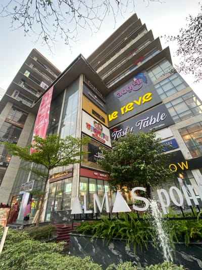 This image presents a low-angle exterior view of the multi-story Almas Point commercial building located in Narayanganj, Bangladesh. The building features a contemporary architectural style with a facade composed of grey concrete and glass panels. Numerous illuminated and non-illuminated signs for retail and restaurant establishments are affixed to the building, including "le reve," "The Tasty Table," "Shoe world," "Priyojon," "ILLIYAN," "AL HASANAT," "WIMBRESSE," "NAYOMI," and "ColourFEST." Bengali script signage includes "বদবু" (Bodhu) and "বাবুবাজার" (Babubazar). A large red vertical banner on the left side displays partial text "KHALERLAN..." and "ALMAS POINT."

At the ground level, a landscaped area is present, featuring green foliage, including ferns, and two small deciduous trees with green leaves. A large, white dimensional sign spelling "ALMAS POINT" is partially visible within this area, with a functional water jet feature obscuring part of the "P" and "O." The base of the landscaped structure is clad in a dark, veined material resembling black marble. Red-carpeted steps lead to a building entrance on the left, where two indistinct figures, one in red attire, are partially visible near a glass storefront. The sky above is light, showing a few bare tree branches at the image's top edge.