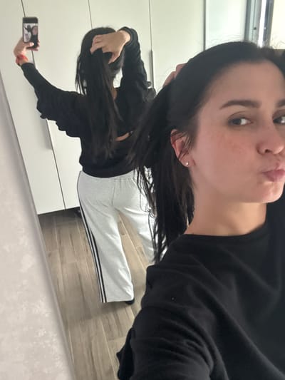 A young woman with long dark hair is taking a mirror selfie in what appears to be a bedroom or dressing area. She is wearing a black long-sleeved shirt and light grey sweatpants with black stripes down the sides. Her hair is partially pulled up, and she is holding a mobile phone in her left hand. The background shows a large white closet with mirrored doors and a wooden floor. The overall lighting is bright and appears to be natural or from overhead fixtures.