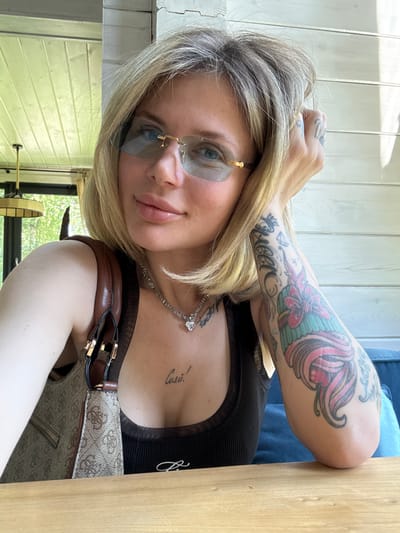 The image displays a close-up portrait of a young woman with blonde, layered hair, captured in a bright, natural-light setting, potentially in Ozernoe, Russia. She is wearing rimless sunglasses with light blue tinted lenses and gold-toned accents. Her expression is calm and confident as she looks directly at the camera, with full lips.

She is dressed in a black ribbed top, possibly a tank top or camisole, featuring a sheer mesh detail at the neckline. A silver-toned necklace with a heart-shaped pendant is visible around her neck. Her left arm is heavily tattooed, featuring a large, colorful design of what appears to be a mermaid or mythical creature with pink and teal elements. Below this, a text tattoo spells out "Gucci" accompanied by a crown and a diamond motif. Smaller tattoos are also visible on her fingers. On her chest, below the collarbone, a script tattoo reads "Силы!", which translates to "Strength!" in Russian.

The woman is leaning on a light-colored wooden table or surface. A brown shoulder bag with a repeating pattern and leather accents is seen next to her. The background includes a light-colored wooden slatted wall and, through a window, lush green foliage and a wooden structure with a rope, suggesting an outdoor or semi-outdoor environment during daytime. The activity is a posed photograph or selfie in a casual, relaxed atmosphere.