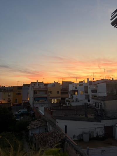 This is an eye-level, outdoor, full shot of a residential cityscape at sunset in Vilanova i la Geltrú, Spain. The sky is a gradient of light blue at the top, transitioning to soft orange and pink hues as it nears the horizon, with scattered wispy clouds. The foreground features a cluster of multi-story buildings, predominantly white and yellow, with tiled roofs. Many buildings have antennas on their rooftops. Some windows are illuminated, suggesting occupancy. In the middle ground, a dark silhouette of a cat is visible on a white wall. To the left, a dark green van is parked near some overgrown vegetation. The image captures a typical urban residential setting bathed in the warm light of twilight.