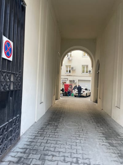 The image captures a view through a long, cream-colored archway in an urban setting, likely in Kraków, Poland. The passageway's floor is paved with grey interlocking concrete blocks.
Through an arched opening at the far end of the passageway, an exterior scene unfolds. A light-colored building with multiple rectangular windows and several air conditioning units is visible. In the open space beyond, several individuals are present: a person in a dark jacket leans against a white car parked on the right, while a red vehicle, possibly a food stall or small truck, stands in the center with more people gathered around it, suggesting a social interaction or transaction.
In the immediate foreground, at the bottom of the image, a black metal grate is visible. Resting on the ground next to it is a "No Stopping" sign, easily identifiable by its blue circular background with a red diagonal cross. The scene appears to take place during daytime, under natural, possibly diffused, light.