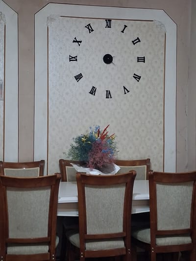The image depicts a wall clock with Roman numerals and a bouquet of dried flowers. The clock face has Roman numerals I through XII, with the hands pointing to approximately 1:40. The clock is set against a patterned wallpaper. To the right of the clock, there are several wooden chairs with upholstered seats, stacked or arranged closely together. A bouquet of dried flowers, featuring a mix of red, blue, and pink hues, is placed near the clock and the chairs. The overall scene appears to be indoors, possibly in a dining area or a room in a house or establishment. The lighting suggests daytime, but no specific time of day can be definitively determined beyond what the clock indicates. There are no people visible in the image. The setting is static, with no discernible activity taking place. The image is likely taken in Yeghegnadzor, Armenia, as per the location context provided. No visible text is present in the image.